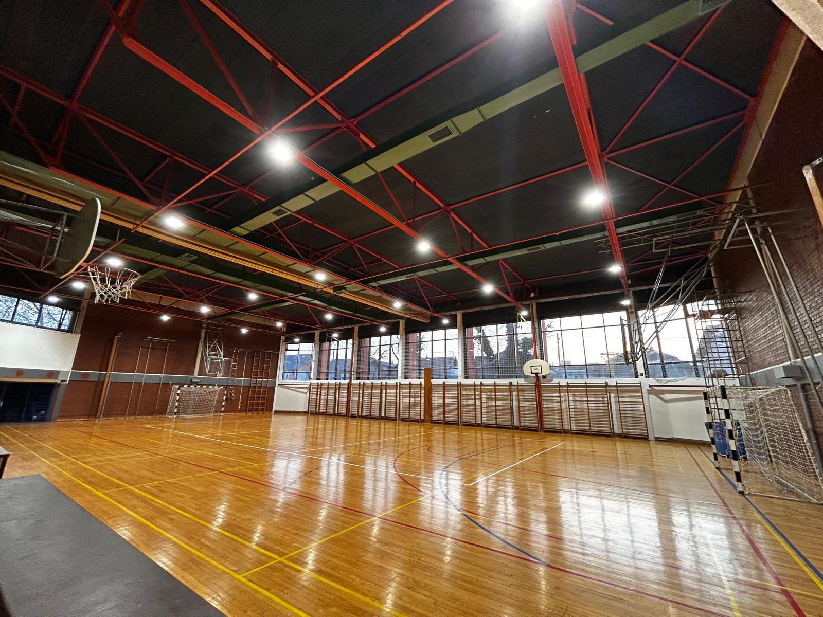 Prostrana teretana s poliranim drvenim podovima, visokim crvenim stropnim gredama, košarkaškim koševima i prozorima koji propuštaju prirodno svjetlo, stvarajući privlačnu atmosferu.