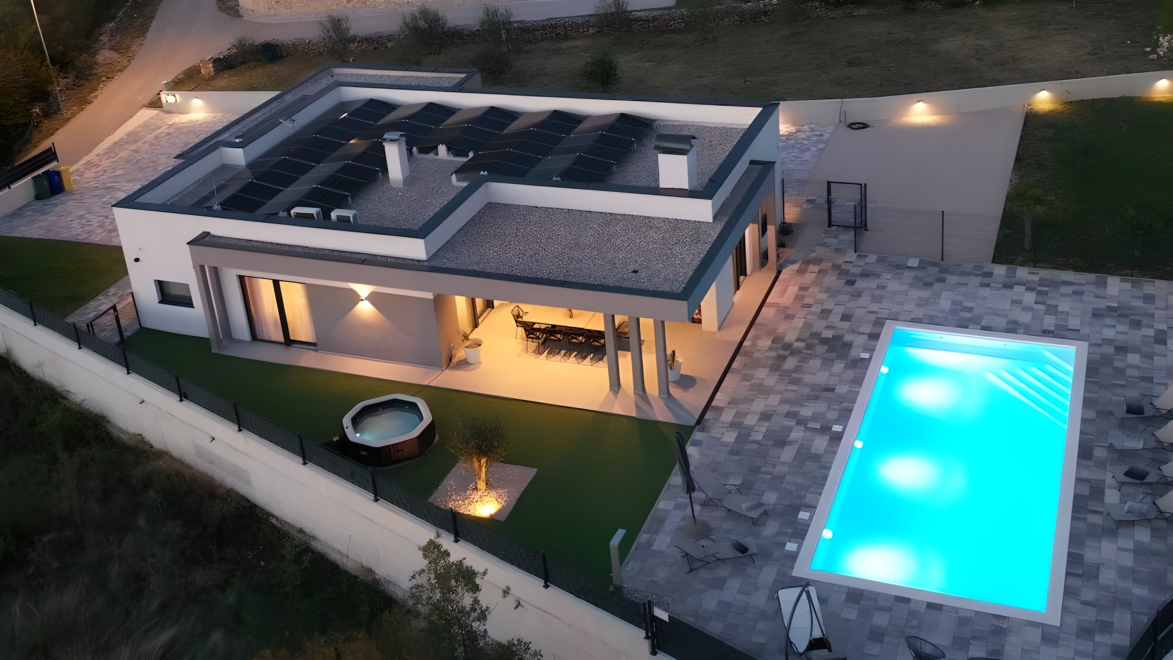 Moderna kuća sa solarnim panelima i osvijetljenim bazenom u sumrak. Terasa s blagovaonicom i okruglim hidromasažnim bazenom. Topla, privlačna atmosfera.