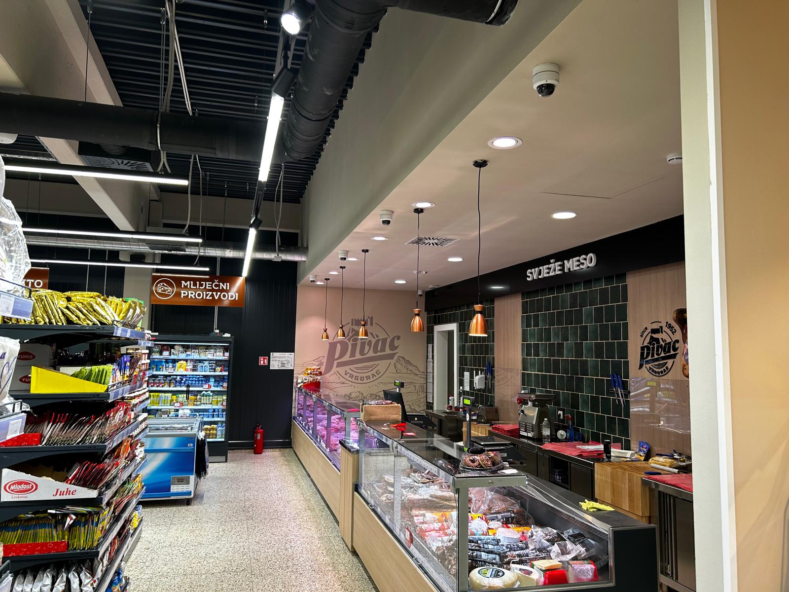 Prolaz supermarketa s policama grickalica i mliječnih proizvoda s lijeve strane i pultom sa svježim mesom i visećim lampama s desne strane. Atmosfera je svijetla i organizirana.