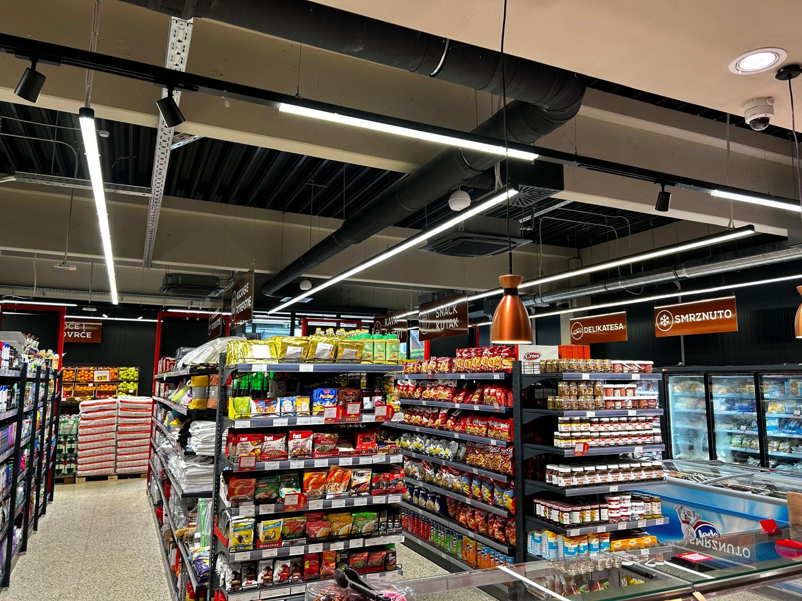 Jarko osvijetljeni prolaz supermarketa s policama punim šarene pakirane robe i grickalica. U pozadini su vidljivi stropni natpisi i zamrzivači.
