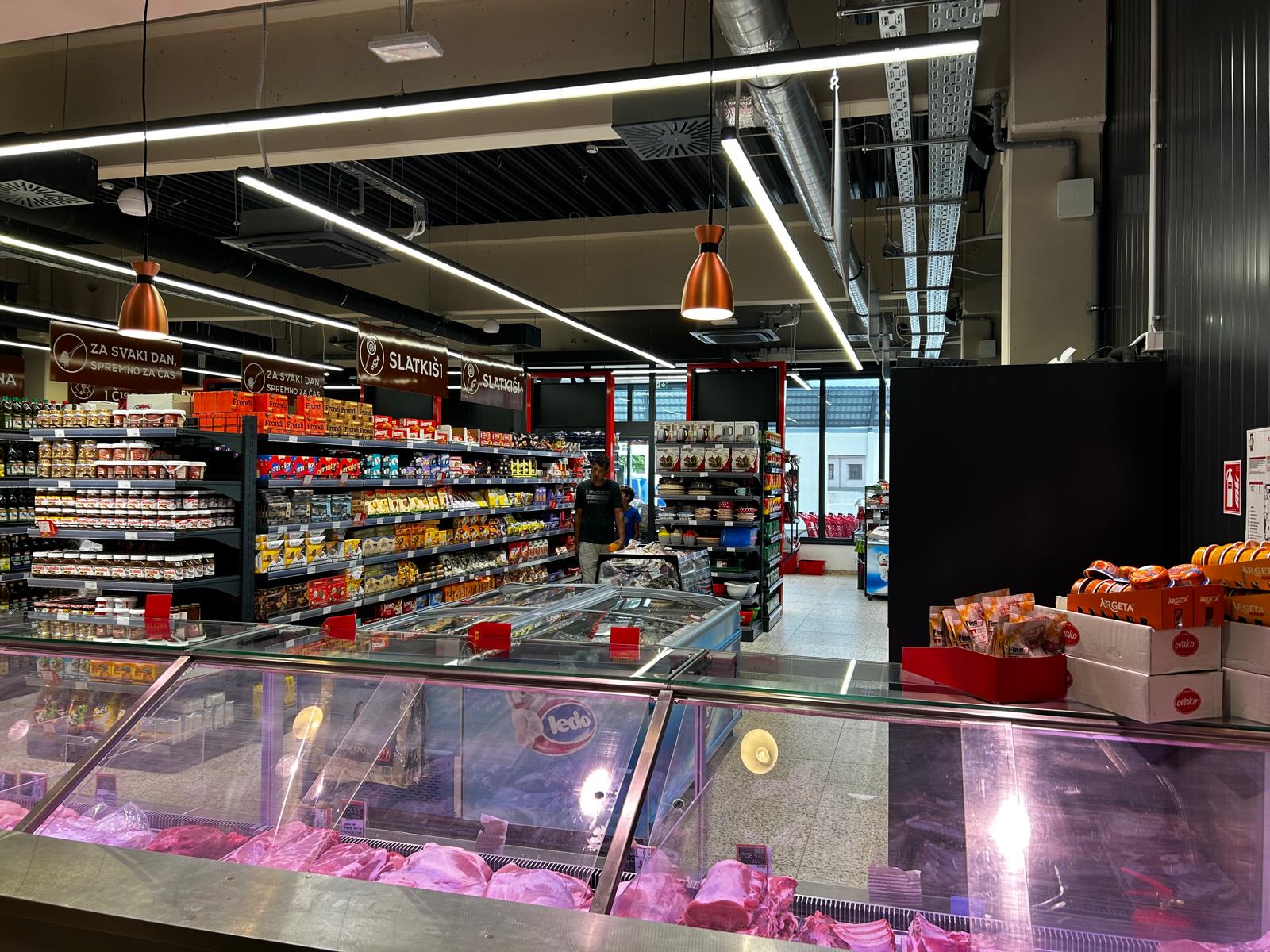 Svijetli interijer supermarketa s rashlađenim vitrinama mesa u prvom planu. Police pune raznih namirnica nižu se u sredini prizemlja. Moderno, čisto i organizirano.