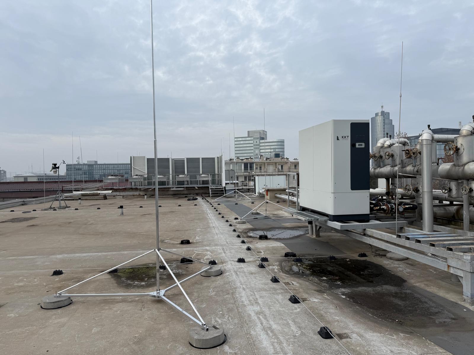 Krov s industrijskom opremom, uključujući antene i cijevi. Oblačno nebo i gradski pejzaž u pozadini stvaraju urbani, industrijski ugođaj.