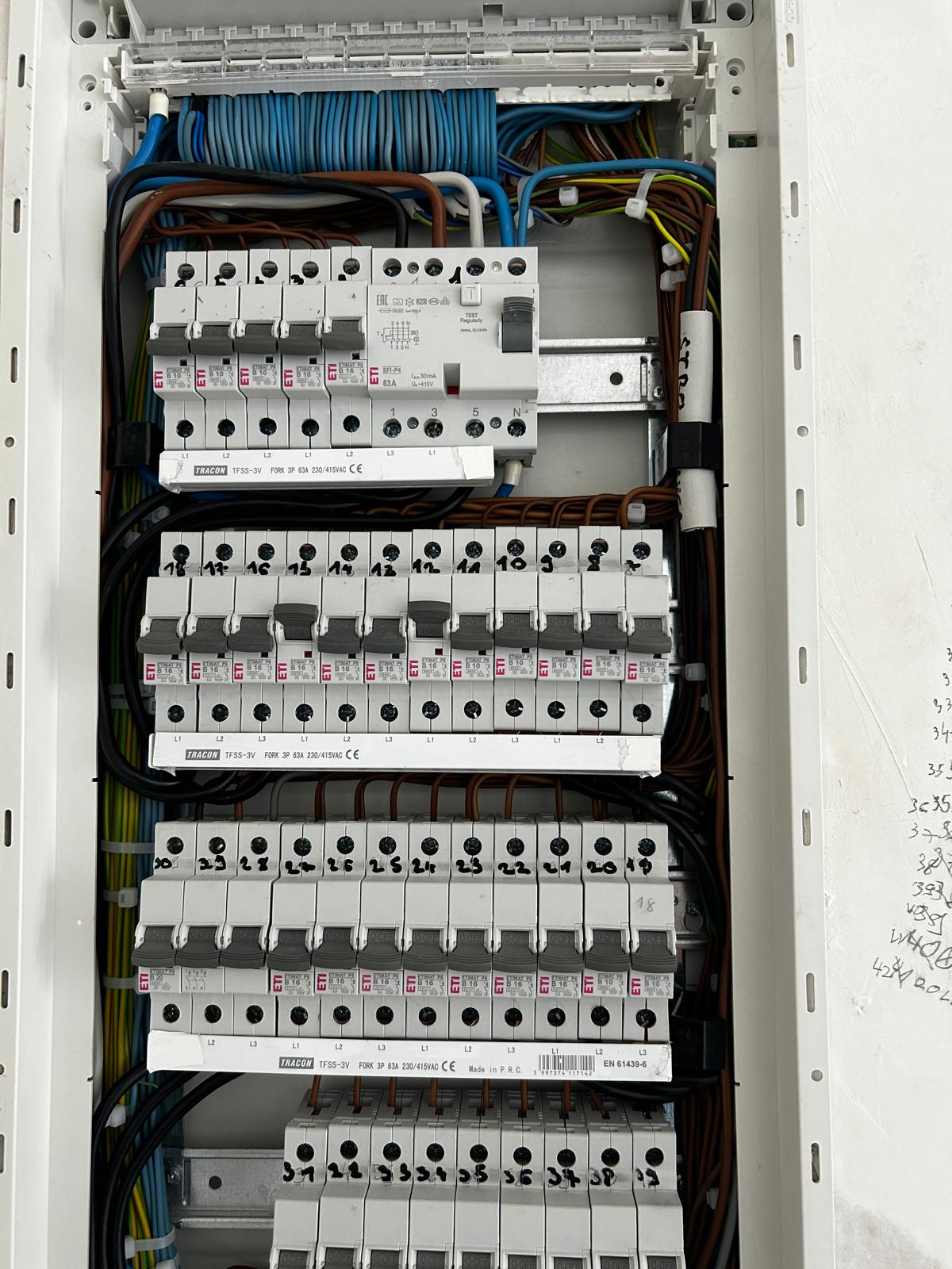 Otvorena električna ploča s više redova označenih prekidača, uredno posloženim žicama u raznim bojama i rukom pisanim bilješkama s desne strane.