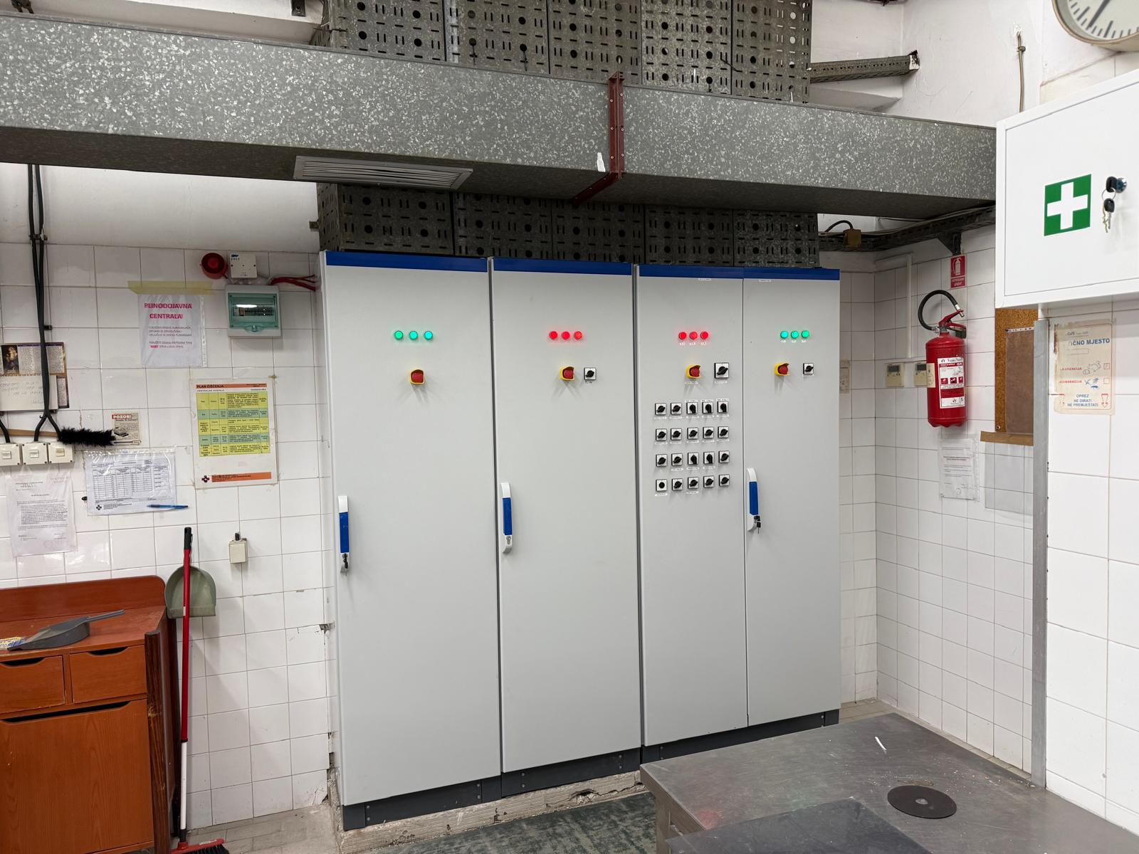 Soba s velikim sivim električnim ormarićima, kontrolnim gumbima i indikatorskim svjetlima. Aparat za gašenje požara i pribor za prvu pomoć vidljivi su na popločanim zidovima.